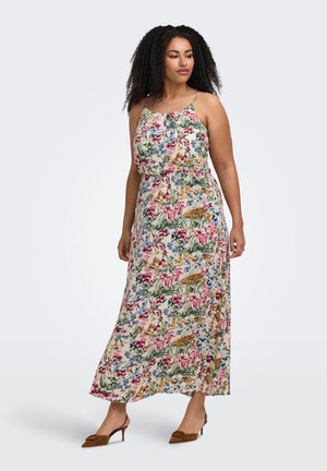 Femme aux cheveux bouclés portant une robe maxi sans manches à motif floral et des talons marron, debout devant un fond clair uni.