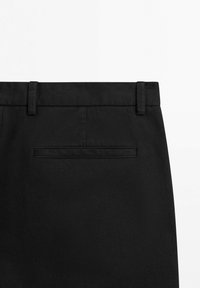 Pantaloni in tessuto nero con tasca a filetto posteriore e passanti per cintura visibili su sfondo bianco, mostrando la parte superiore della schiena.