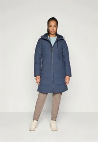 Žena stoji u dugom tamnoplavom kaputu s kapuljačom, svijetloplavoj košulji, bež hlačama i bijelim tenisicama, naslonjena na jednostavnu pozadinu.