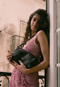 Černá texturovaná kabelka s jedním ramínkem, držená na růžových šatech s volánkovými detaily. Na přední straně je zlaté reliéfní logo.