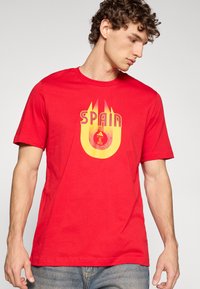 Červené bavlněné tričko s žlutým potiskem "ŠPANĚLSKO" a emblémem uprostřed. Krátké rukávy a kulatý výstřih.