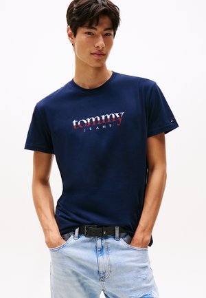 Jeune homme portant un t-shirt Tommy Jeans bleu marine et un jean déchiré bleu clair, mains dans les poches devant un fond uni.