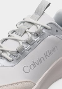 Λεπτομέρεια λευκού αθλητικού παπουτσιού Calvin Klein που δείχνει τα κορδόνια να περνούν μέσα από γκρι υφασμάτινες θηλιές και ανάγλυφο το όνομα της μάρκας στο πλευρό.