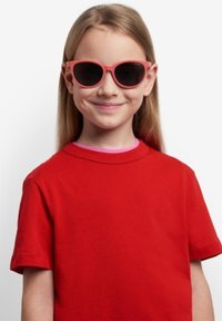 Occhiali da sole rosa con lenti scure su un bambino. Indossa una maglietta di cotone rossa, a maniche corte, e ha i capelli lisci. Sfondo semplice.