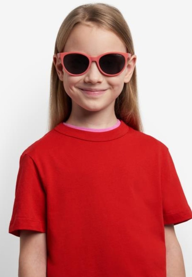 Occhiali da sole rosa con lenti scure su un bambino. Indossa una maglietta di cotone rossa, a maniche corte, e ha i capelli lisci. Sfondo semplice.