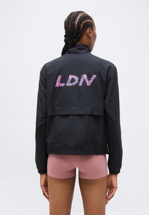 Persona con capelli intrecciati che indossa una giacca nera con la scritta "LDN" in motivi rosa sulla schiena e pantaloncini rosa, vista di spalle.