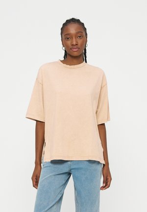 Νεαρή γυναίκα με πλεγμένα μαλλιά όρθια, φοράει μπεζ φαρδιά κοντομάνικη μπλούζα και ανοιχτό μπλε τζιν μπροστά σε απλό φόντο.