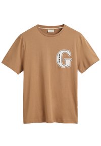 Camiseta de algodón color marrón claro con cuello redondo, mangas cortas y una gran letra blanca "G" con la palabra "GANT" impresa en negro debajo.