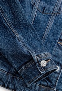 Rękaw kurtki dżinsowej złożony na ciele, z widocznymi szwami i metalowym guzikiem z grawerunkiem "NEXT JEANS SINCE 1982."