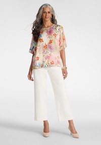 Mujer con cabello largo y gris, vestida con una blusa floral colorida, pantalones anchos blancos, tacones nude y pulseras doradas, sobre un fondo liso.