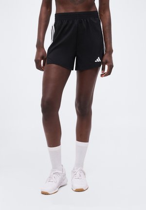 Άτομο που φοράει μαύρα σορτς Adidas με λευκές ρίγες, λευκές κάλτσες και άσπρα αθλητικά παπούτσια, στέκεται μπροστά σε απλό φόντο.