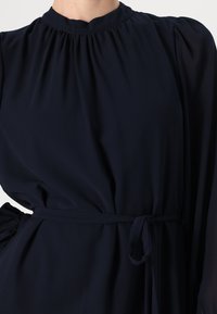 Tamno plava haljina dugih rukava od lagane tkanine s visokim izrezom, s nabranim detaljima i vezanom strukom za uski siluetu.
