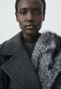 Cappotto grigio in lana con un ampio colletto, caratterizzato da un accento in pelliccia sintetica grigia strutturata sulla spalla. Tessuto liscio, design strutturato e look moderno.