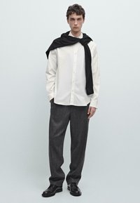 Camicia bianca con bottoni, pantaloni grigi texturizzati, maglione nero appoggiato sulle spalle, scarpe nere. Sfondo neutro.