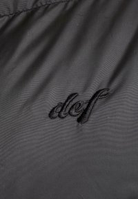 Sort stof har en glat tekstur med et broderet "def" logo i matchende sort tråd. Designet er minimalistisk og moderne.