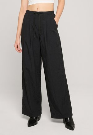 Kvinde iført sort vidde bukser med høj talje og snøre i taljen, sidelommer, kombineret med sorte hælede støvler og en hvid cropped top.