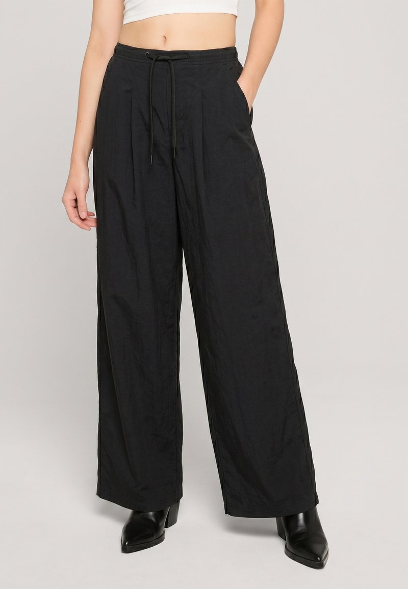 Femme portant un pantalon noir taille haute à jambes larges avec ceinture à cordon, poches latérales, associé à des bottines noires à talons et un crop top blanc.
