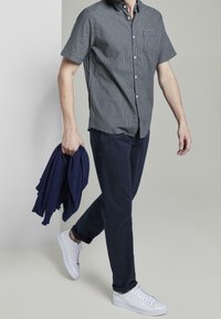Camicetta a maniche corte con motivo in colori scuri, pantaloni blu navy e sneakers bianche. Tiene una sciarpa blu navy. Tessuto morbido, vestibilità comoda.