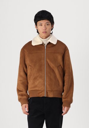 Ruda zomšinė bomber tipo striukė su balta avikailio apykakle, gumbais apykaklėje ir apačioje, priekinis užtrauktukas ir dvi šoninės kišenės.