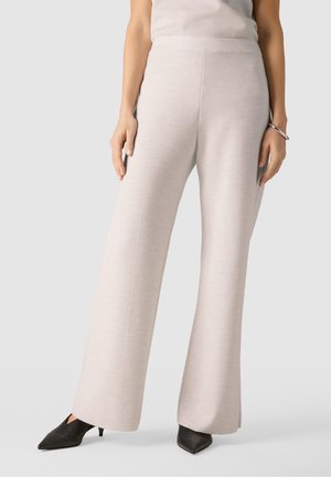 Kobieta w jasnobeżowych, szerokich spodniach w połączeniu z czarnymi szpilkami w szpic, stojąca na tle jednolitego jasnego tła.