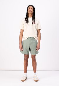T-shirt léger beige à manches courtes avec un graphique rouge, associé à un short vert olive et des chaussures blanches, se tenant sur un fond uni.