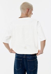 Bílé oversized tričko s embosovaným geometrickým vzorem a textem "TY PÍŠEŠ PŘÍBĚH" na zádech. Nosené s tmavě modrými džíny.