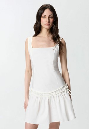 Femme aux longs cheveux foncés portant une robe blanche sans manches avec un col carré et une jupe froncée sur un fond clair uni.