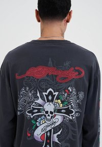 Schwarz langärmliges Shirt mit einem bunten Schädel- und Knochen-Design auf dem Rücken, mit kontrastierendem roten Text und filigranen floralen Mustern.