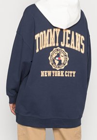 Námořnická mikina s bílou kapucí, s velkým logem "TOMMY JEANS" a designem globu na zadní části. Doplňující text "NEW YORK CITY" umístěný níže.