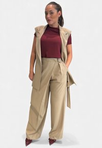 Gilet beige su misura sopra una camicetta bordeaux, abbinato a pantaloni beige a gamba larga. Completa il look un paio di décolleté bordeaux con punta affilata.
