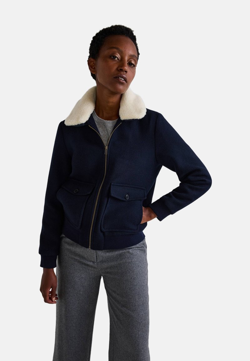 Veste zippée bleu marine avec un col en fausse fourrure crème, deux poches à l'avant, des poignets et un ourlet côtelés, associée à un pantalon gris.