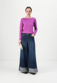 Fialový top s dlhým rukávom skombinovaný s širokými tmavými džínsami s jemnými svetlomodrými manžetami. Modelka drží malú textúrovanú kabelku.