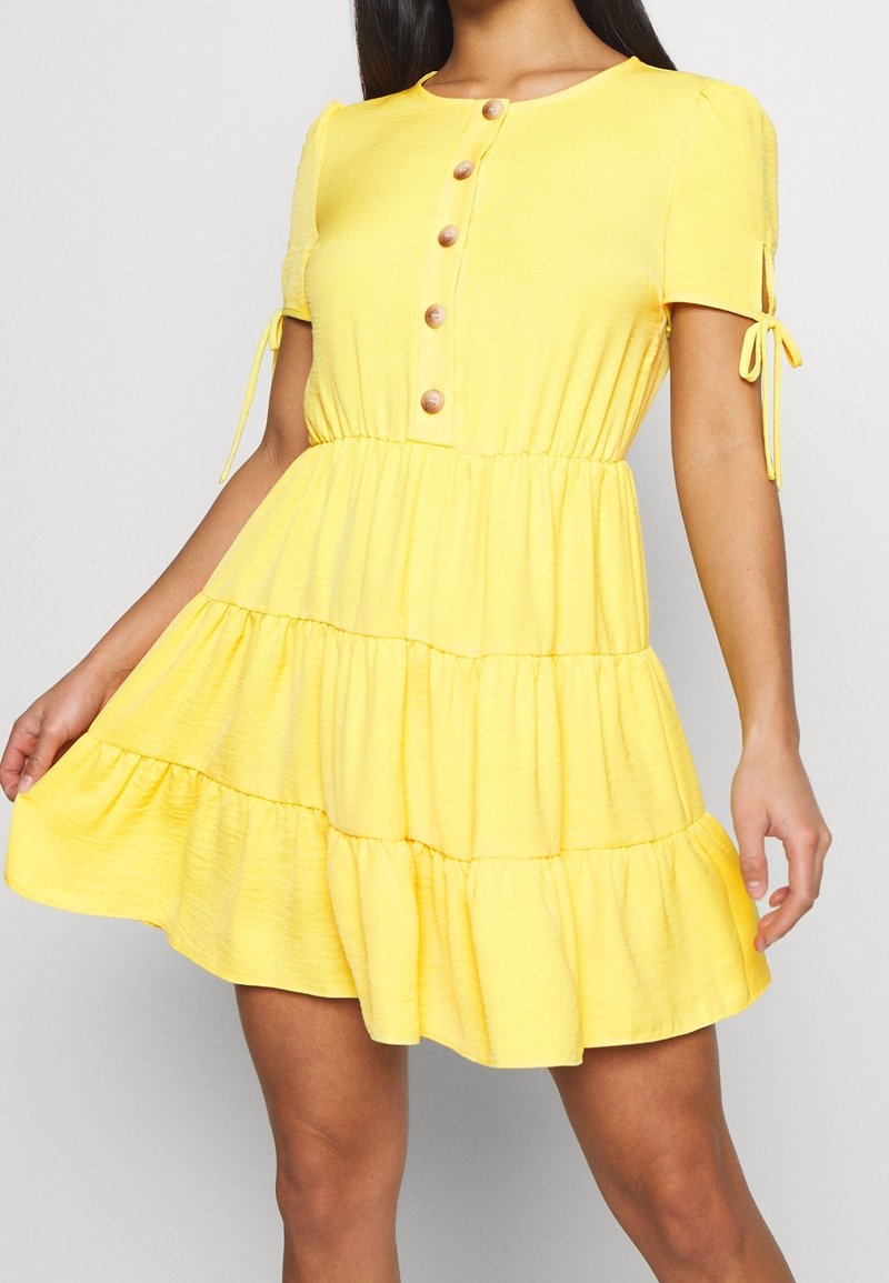 Žluté šaty s volány vyrobené z měkké tkaniny. Mají krátké rukávy, kulatý výstřih a knopky na přední straně. Obsahují ozdoby na rukávech se stahovacími detaily.