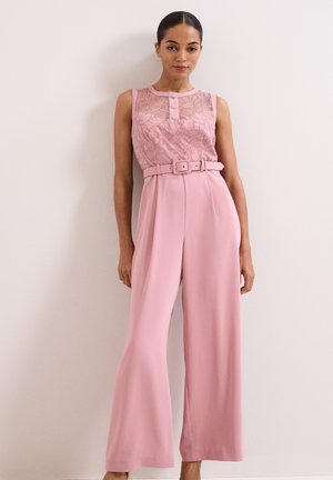 Femme portant une combinaison rose sans manches avec un haut en dentelle, des détails de boutons, une ceinture assortie et un pantalon large, debout contre un mur uni.