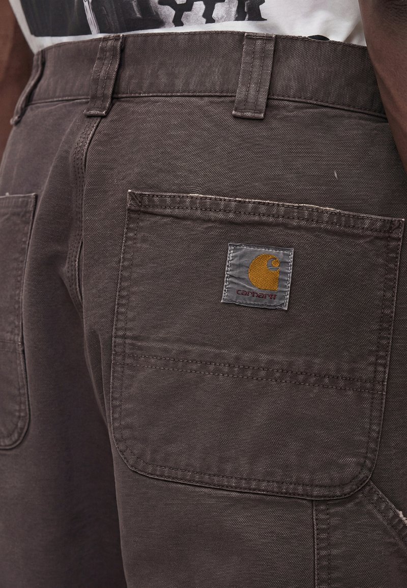 Nahaufnahme einer dunkelbraunen Carhartt-Hose mit Gesäßtasche und Carhartt-Logo-Patch auf der rechten Seite.