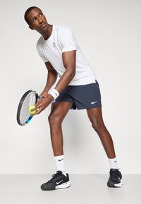 Λευκό αθλητικό μπλουζάκι, ναυτικές σορτς με πλαϊνές τσέπες, μαύρη ρακέτα τένις, πράσινη μπάλα τένις, μαύρα και λευκά αθλητικά παπούτσια, λευκή ποδοσφαιρική ταινία.