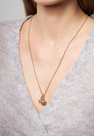 Goldene Kettenhalskette mit einem runden Anhänger, der einen Sternelement und eine strukturierte Oberfläche aufweist, getragen über einem weichen, hellgrauen Pullover.