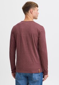 Langærmet maroon T-shirt lavet af blødt stof, der har en rund halsudskæring og et lille mærke nederst til venstre, parret med blå jeans.