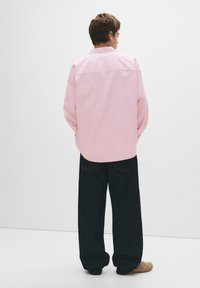 Cămașă roz cu dungi, cu mâneci lungi, margine rotunjită și pantaloni din denim închis. Vederea din spate evidențiază materialul moale și croiala lejeră.