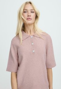 Różowy sweter z mieszanki wełny, krótki rękaw, z prążkowanym wykończeniem u dołu i przy kołnierzu. Z przodu znajdują się trzy dekoracyjne guziki z kwiatowym wzorem.