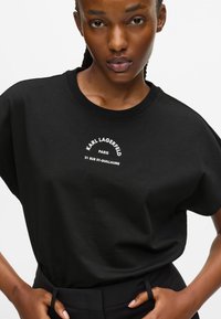 Fekete pamut póló laza szabással, fehér nyomtatott szöveggel: "KARL LAGERFELD PARIS 21 RUE ST-GUILLAUME." Sima textúrájú.