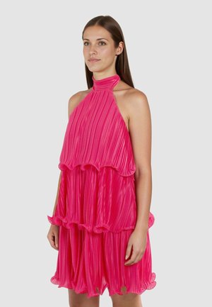 Moteris su tiesiais rudos spalvos plaukais, vilkinti rankovių neturinčią, sluoksniuotą, plisuotą ryškiai rožinę suknelę su aukštu kaklu, stovinti priešais paprastą foną.