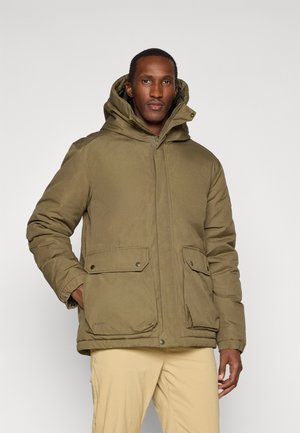 Man wearing an olive green hooded winter jacket with front zipper and flap pockets, paired with light beige pants, standing against a white background.
