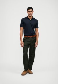 Camisa polo azul marino de manga corta, combinada con pantalones verdes oliva y un cinturón marrón. Unos zapatos marrones completan el conjunto.