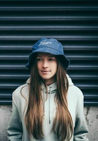 Denim bucket-hattu, jossa on sininen väri ja valkoinen brodeerattu logo, yhdistettynä vaaleanharmaaseen huppariin. Pitkät hiukset näkyvät hatun alta.