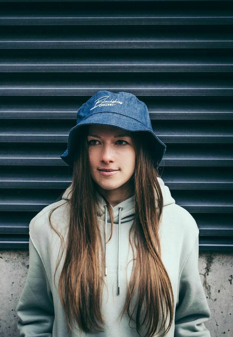 Denim bucket-hattu, jossa on sininen väri ja valkoinen brodeerattu logo, yhdistettynä vaaleanharmaaseen huppariin. Pitkät hiukset näkyvät hatun alta.