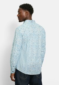 Helles blaues Hemd mit langen Ärmeln und einem kleinen, verstreuten Muster aus dunkelblauen Formen. Klassischer Kragen, entspannte Passform und Knopfmanschetten.