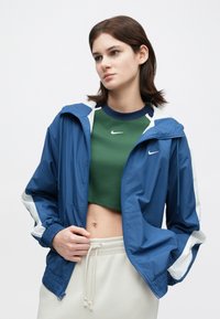 Nő kék és fehér zipzáras dzsekiben, zöld crop top fölött, világos bézs melegítőnadrágban, egy sima háttér előtt áll.