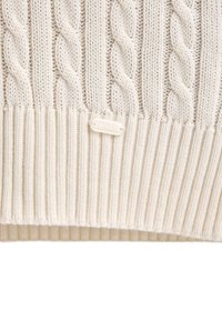 Beiger Strickpullover mit Kabelmuster, der einen gerippten Saum und ein kleines Logoetikett mit der Aufschrift "POLO CLUB" am unteren Rand aufweist. Weiche Textur und grobes Design.