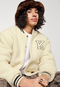 Cream shearling bomber jacket with black stripes at the cuffs and collar, featuring letter "K" patch. Brown fur bucket hat. Plaid pants.